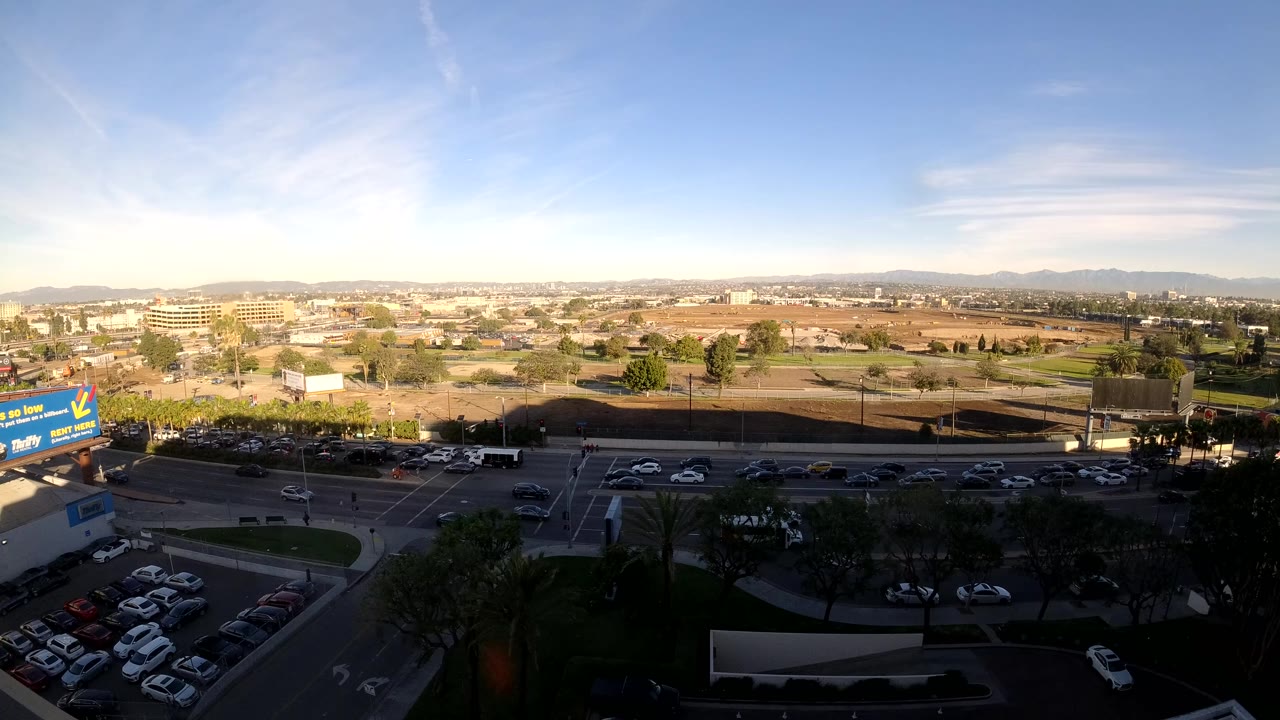 Opening frame of Los Angeles traffic from an elevated viewpoint, with cars on a multi-lane road, a parking lot in the lower left, low buildings, distant hills, and tiny distant people not clearly identifiable.