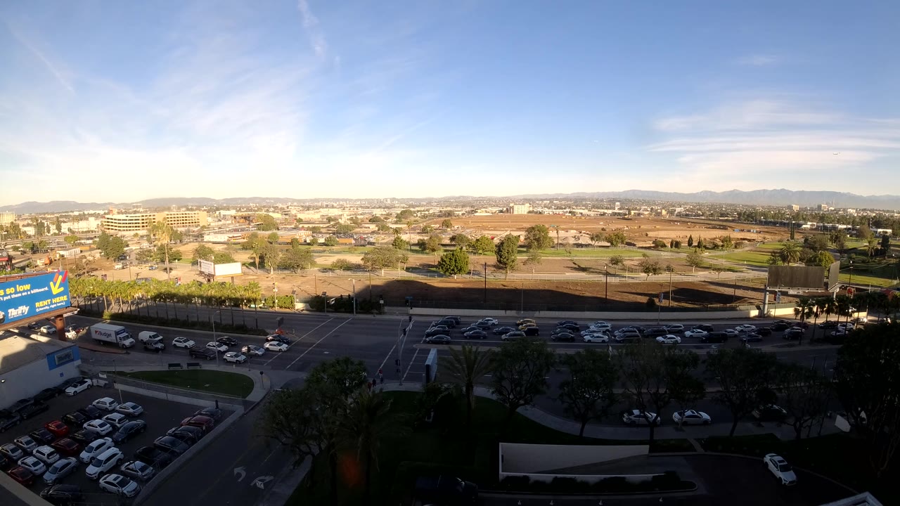 Mid-clip frame showing Los Angeles cars spread along the road, foreground trees in shadow, parked vehicles below, low-rise development in the distance, and a clear blue sky above.