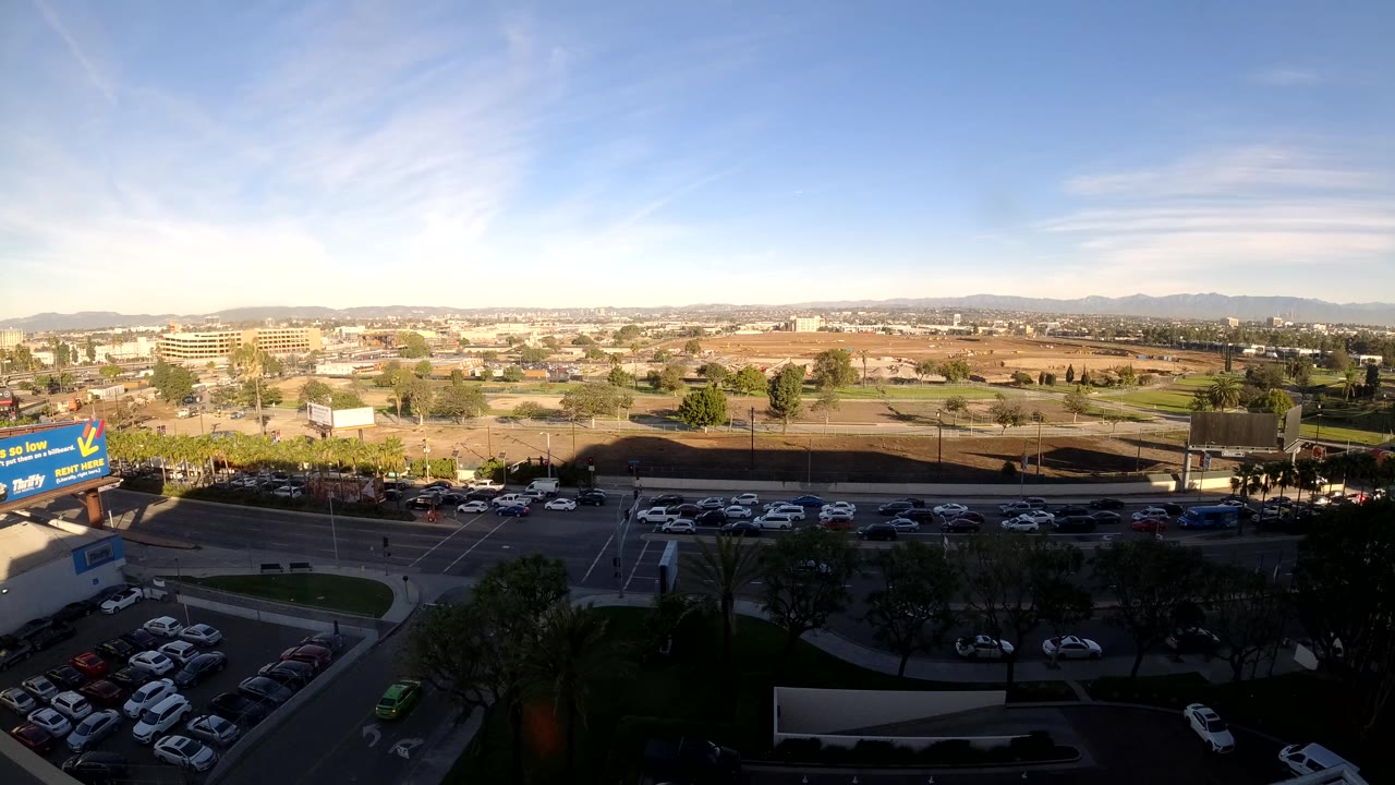 Wide elevated daytime view in Los Angeles, California, with cars moving on a street below, parked vehicles in lots, dark foreground trees, dry open land, low buildings, distant hills, and no visible people.