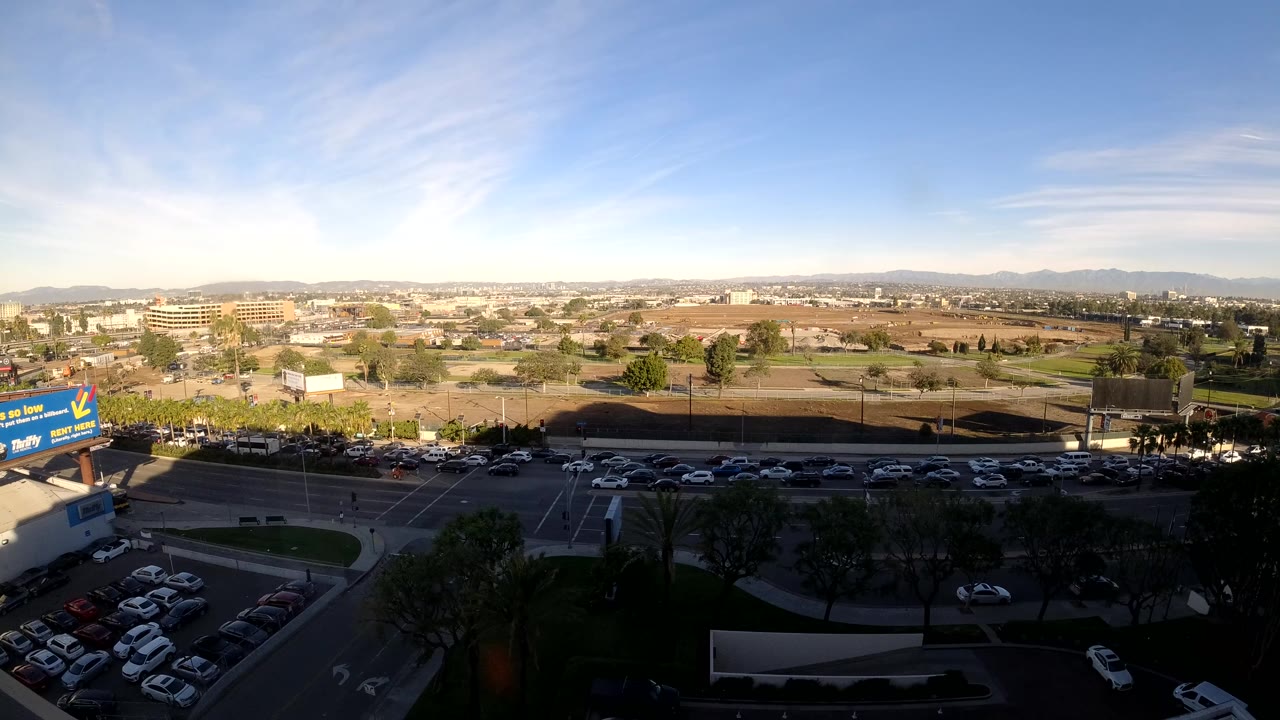 Opening frame of Los Angeles from an elevated view, with parked cars in shadow, traffic on a multi-lane road, low buildings, distant hills, bright blue sky, and no visible people.