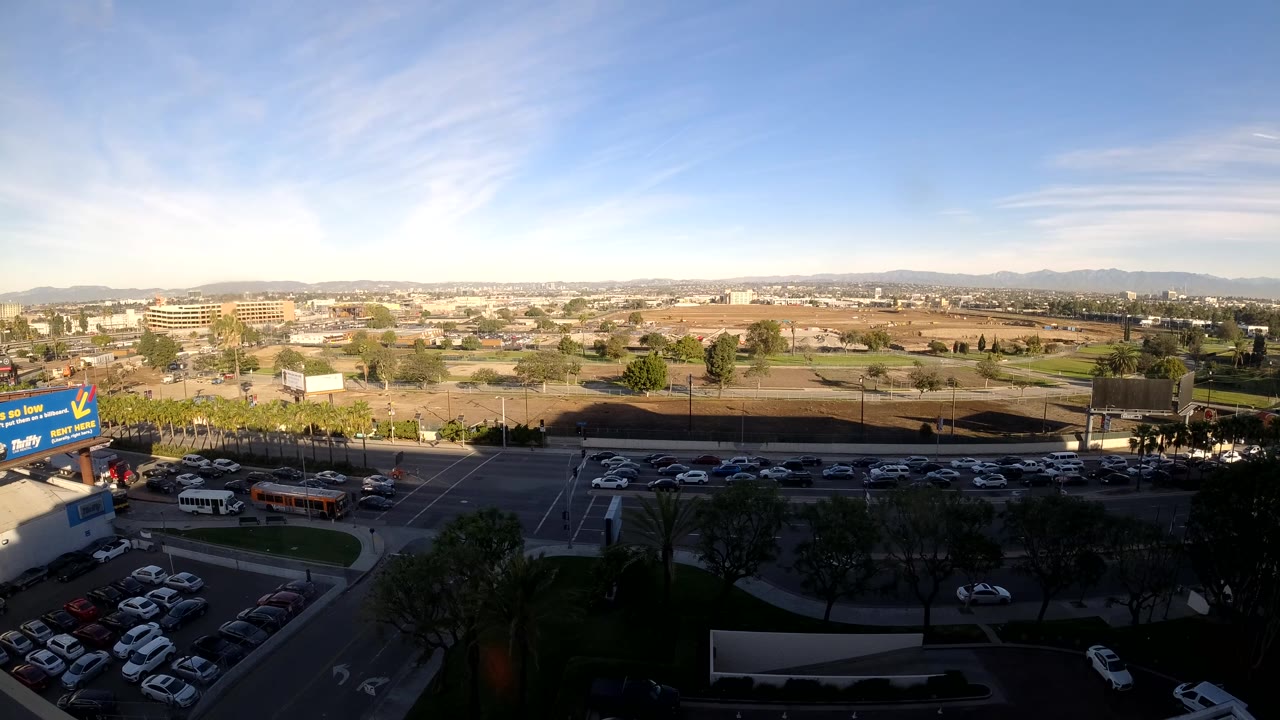 Third frame of the Los Angeles traffic scene, with moving cars on the road, a broad parking area in the lower frame, commercial buildings across the midground, distant hills, and no visible people.