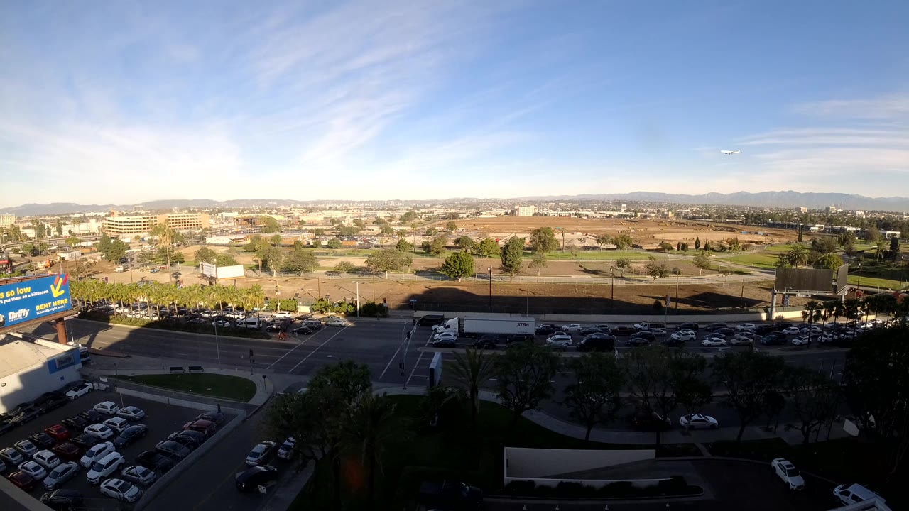 Wide Los Angeles aerial frame showing cars progressing along the lower road, shaded trees in the foreground, commercial lots beyond, distant mountains, and no visible people.