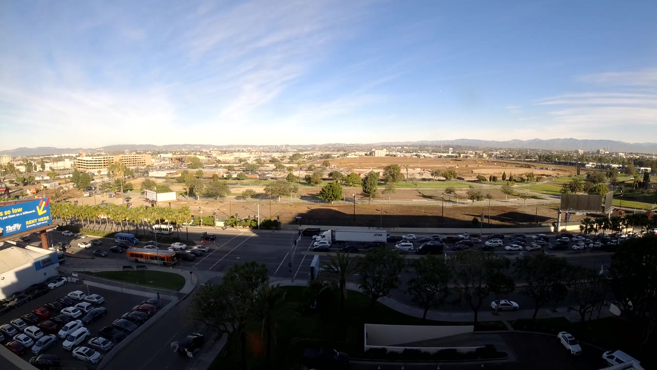 Los Angeles road traffic passes below the camera near a blue billboard, with parking rows, dry open ground, low industrial buildings, distant hills, and no visible people.