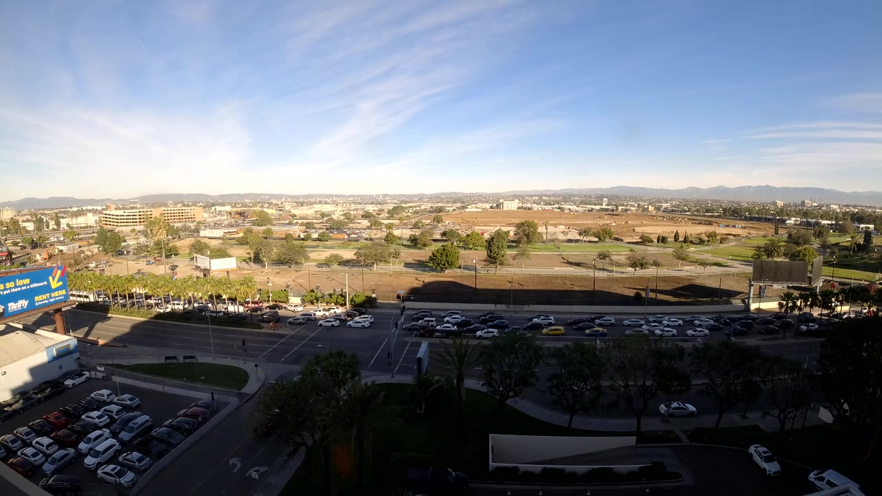 Opening frame of Los Angeles traffic from an elevated view, with cars moving along a broad road, parking lots in shadow, dry open lots, distant hills, blue sky, and no visible people.