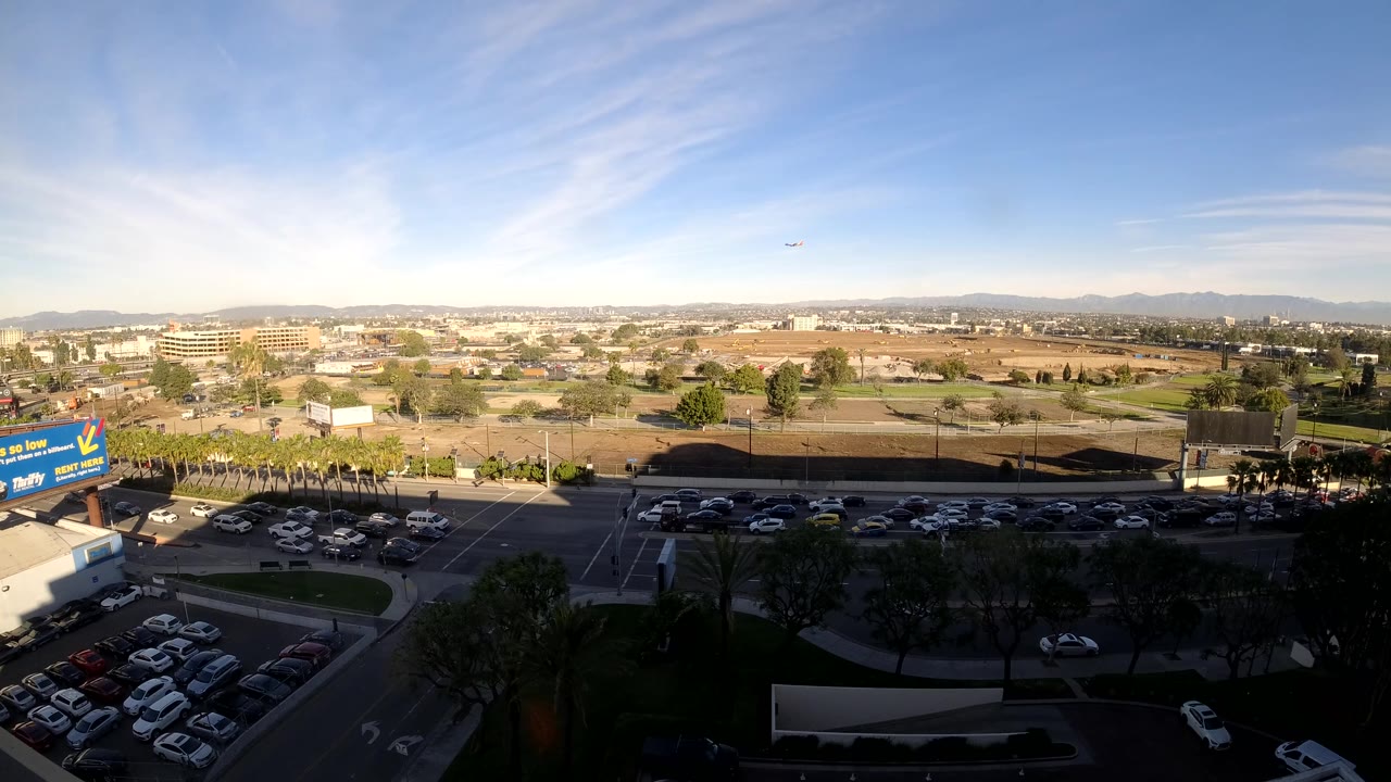At 0:02, the Los Angeles view shows cars and trucks spaced along the road beneath shadowed foreground trees, parking areas, tan open lots, low buildings, distant hills, and no visible people.