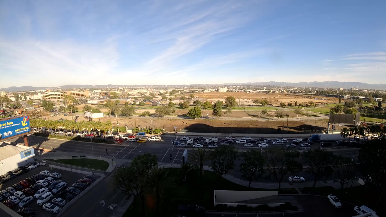 Second frame holds the elevated Los Angeles composition as traffic fills the avenue, with parked cars at lower left, open lots in the middle distance, and no visible people.