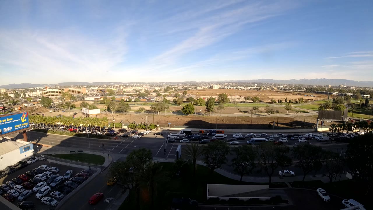 Opening frame of a Los Angeles aerial view with parked cars in the lower left, a busy road across the middle, low buildings beyond, distant hills, and no visible people.