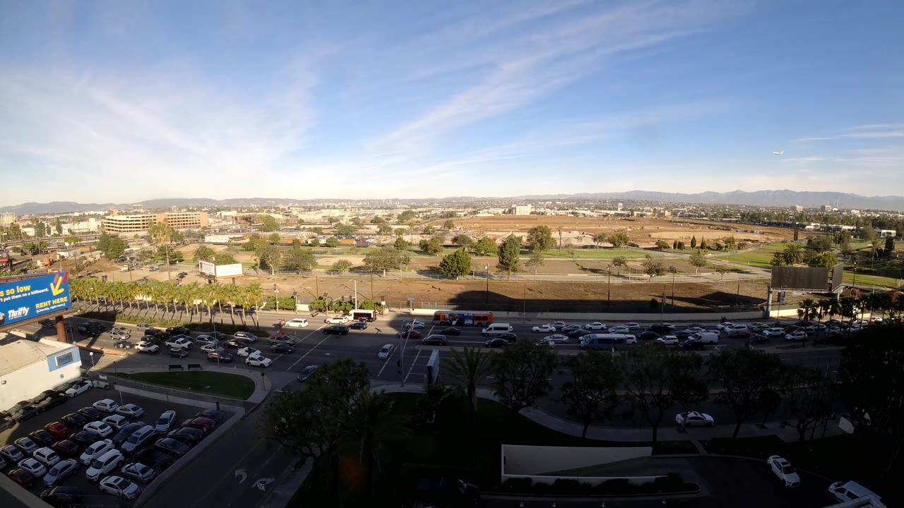 At two seconds, the Los Angeles aerial view shows cars moving along the road, a shadowed foreground lot, trees, open development land, distant mountains, and no visible people.