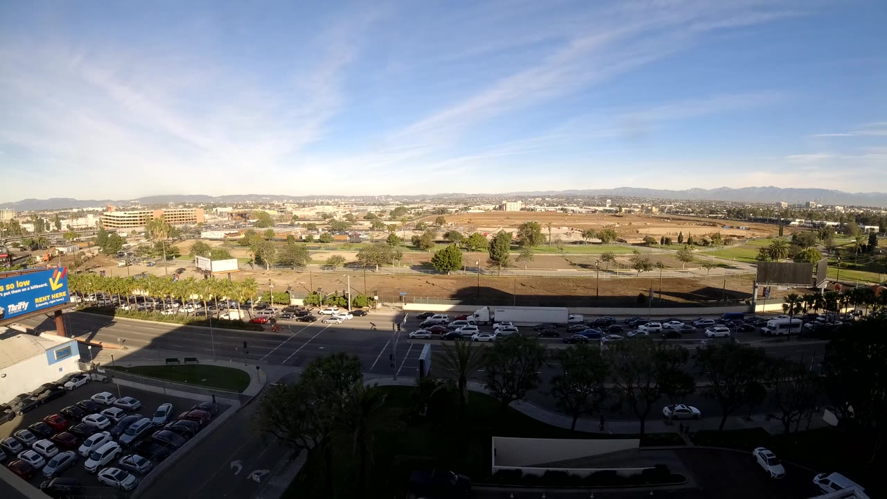 Opening elevated view in Los Angeles showing a busy road with cars, parking lots in shadow, low-rise buildings, open lots, distant hills, and no visible people.
