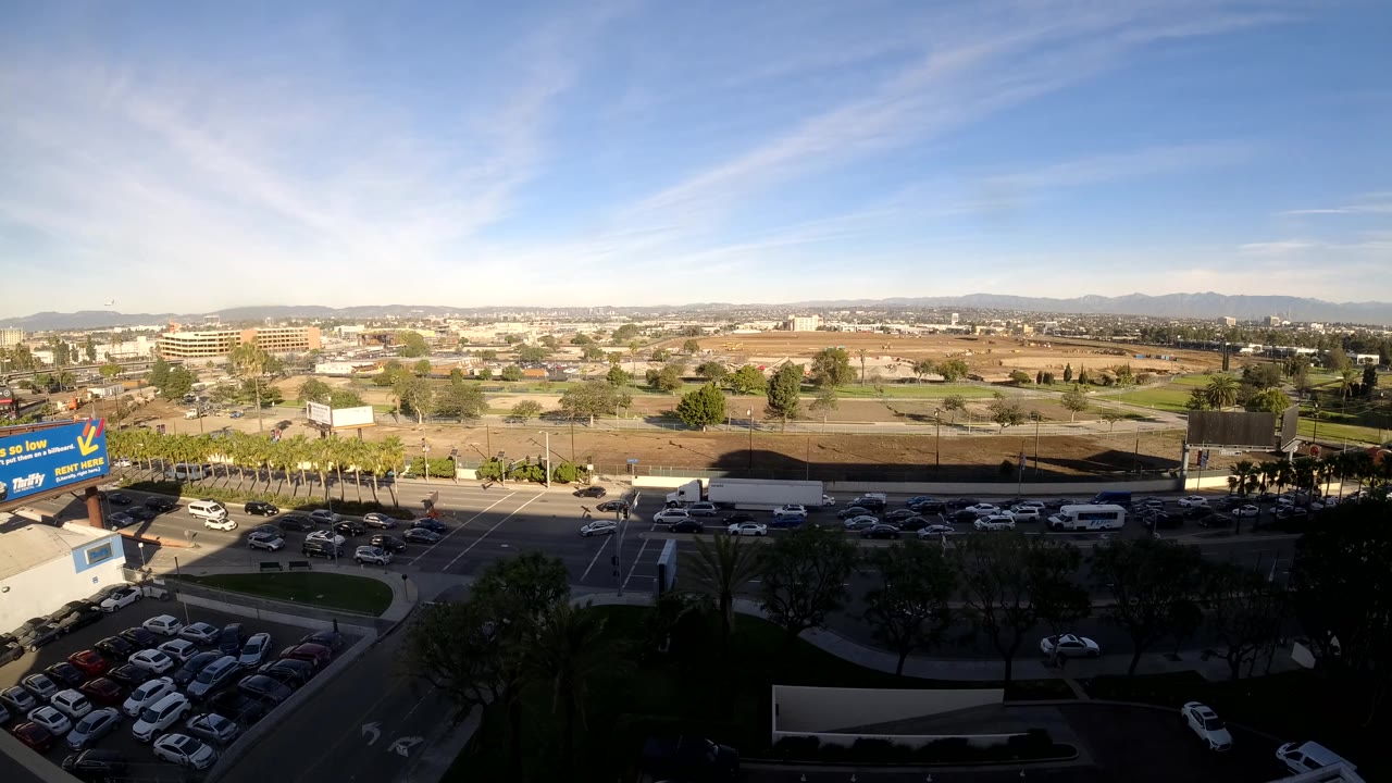 At 0:02, the Los Angeles road remains busy with cars passing in both directions, a white vehicle near the lower road edge, open lots behind, and no visible people.