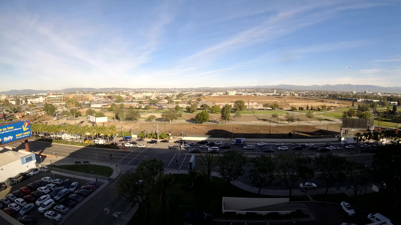 Elevated daytime view of a road full of cars, a parking lot, trees, and dry open land.