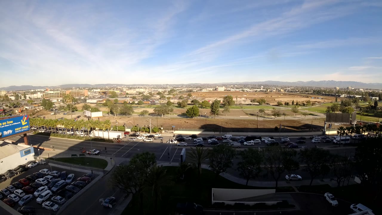 Los Angeles elevated wide view with cars moving along a road, a parking lot in the foreground, low buildings and distant hills beyond, and no visible people.