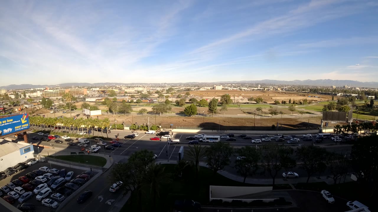 Los Angeles daytime traffic moves past parking areas and open lots, with dark trees at the bottom of frame and low city buildings across the horizon, no visible people.