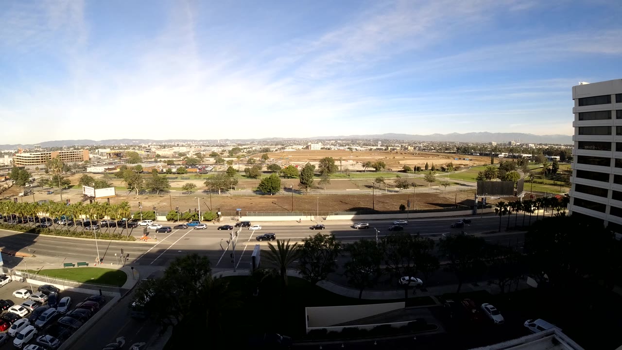 Opening elevated view in Los Angeles showing light road traffic, parked cars, open lots, distant hills, a tall white building on the right, and no visible people.