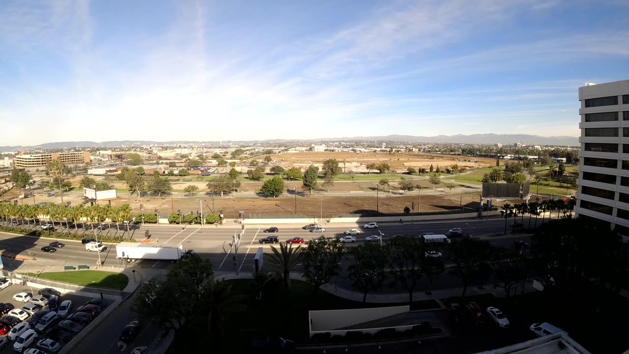 Los Angeles traffic view with a large white truck passing through the road area, smaller cars nearby, open lots in the background, a white tower at right, and no visible people.