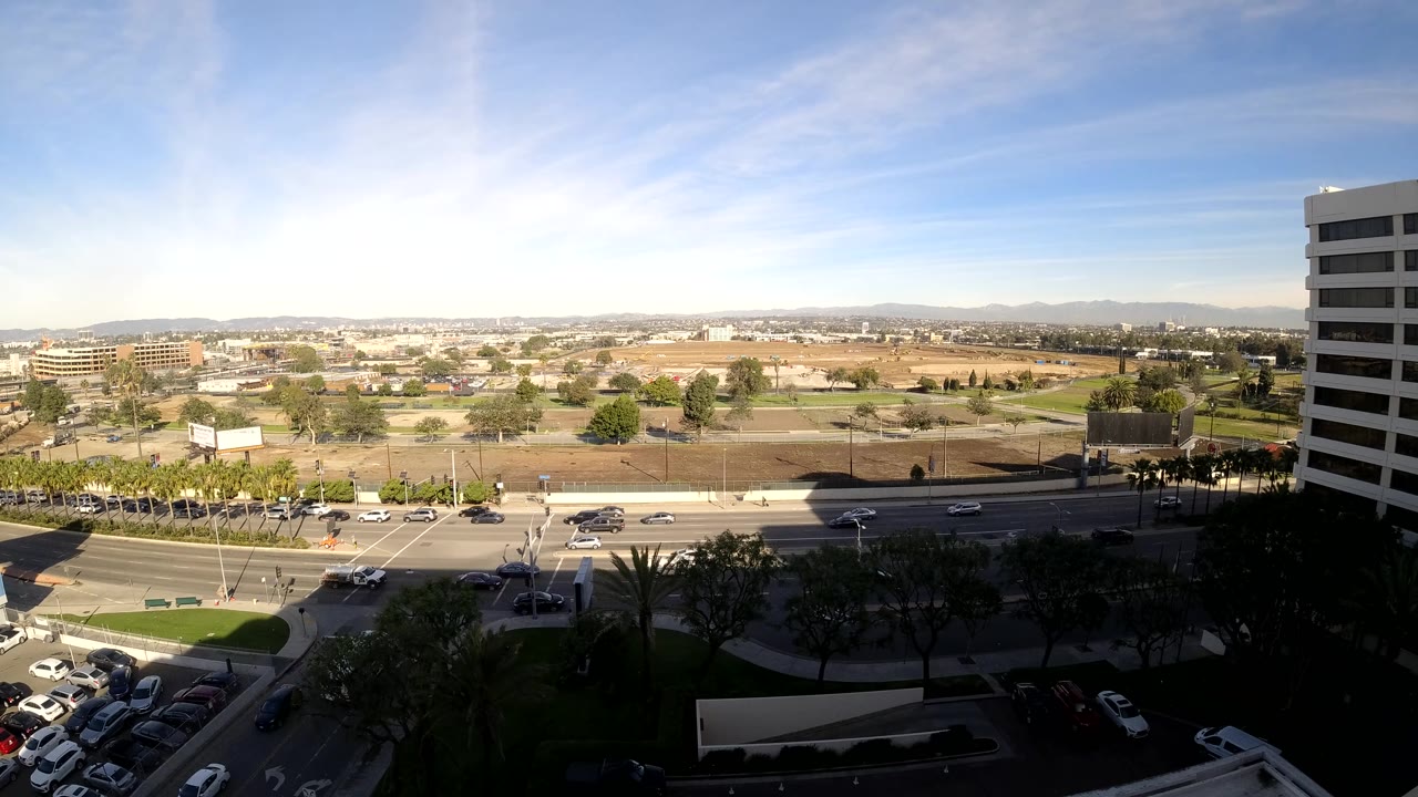 Mid-clip Los Angeles frame with cars spaced along the road, shadowed foreground trees and parking, open lots, low buildings, distant hills, and no visible people.
