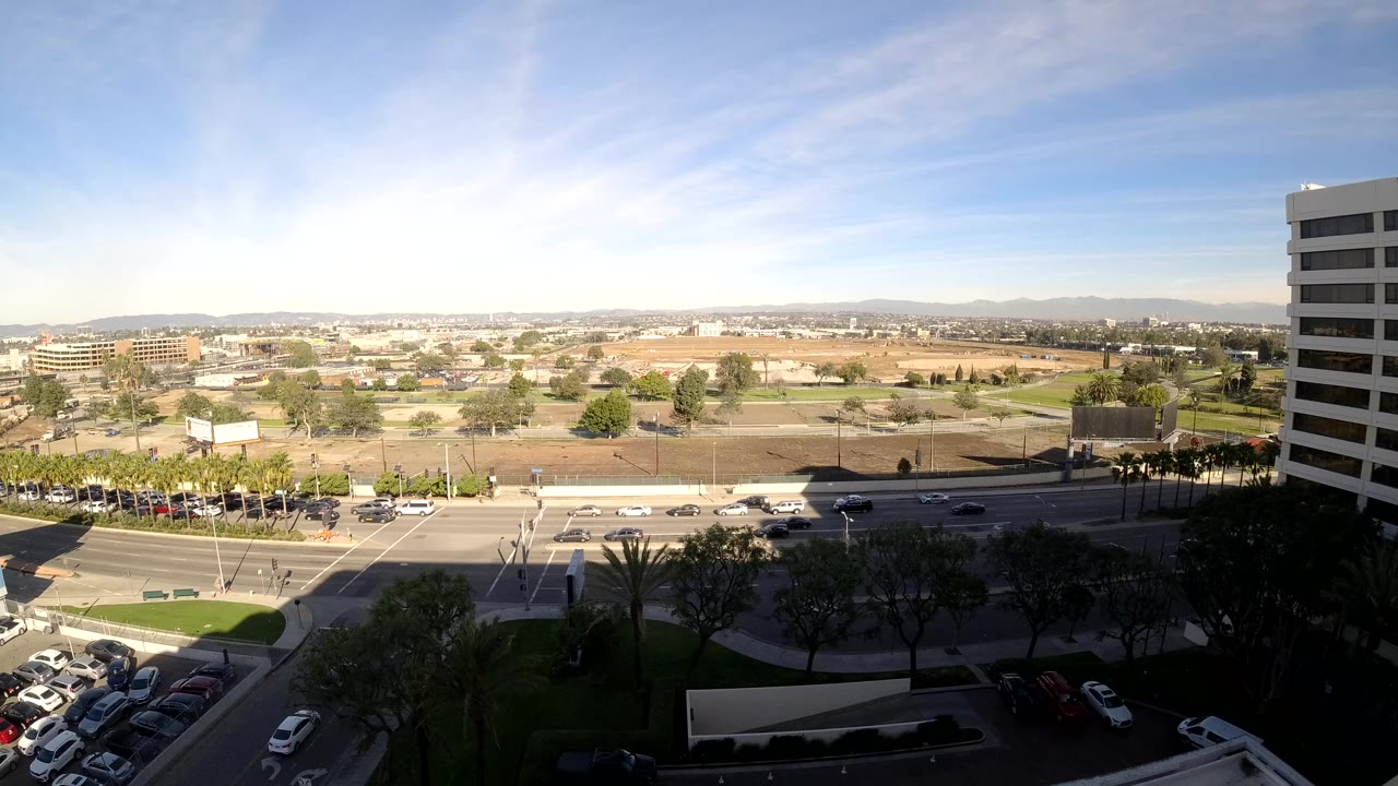Second frame shows the Los Angeles road scene from above, with more cars crossing the midground, parked vehicles at lower left, a white building at right, and no visible people.