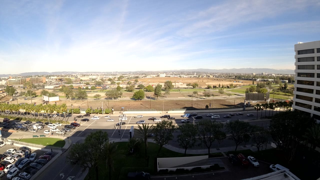 At 0:02, the Los Angeles view includes cars clustered near the lower road area, open land beyond the street, a tall building edge on the right, and no visible people.