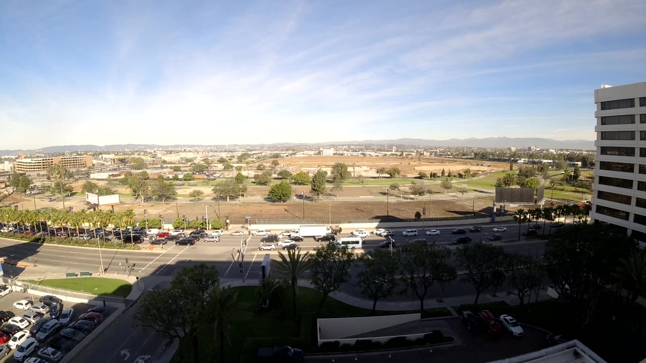 Wide Los Angeles city frame showing road traffic below, a dark landscaped foreground, tan open lots, distant hills, blue sky, and no visible people.