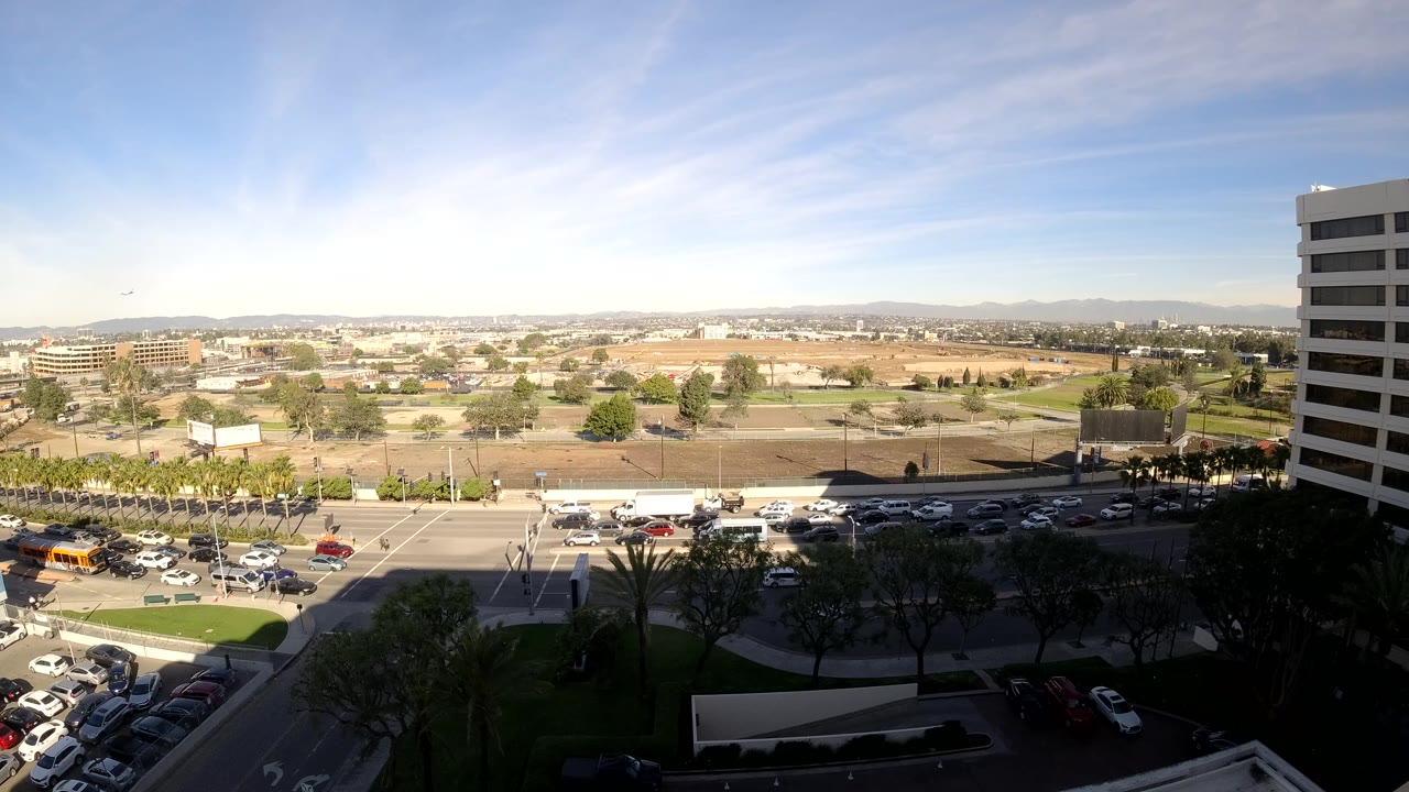 Los Angeles daytime road scene from above with cars, a yellow bus, parking rows, low buildings, distant hills, and no visible people.