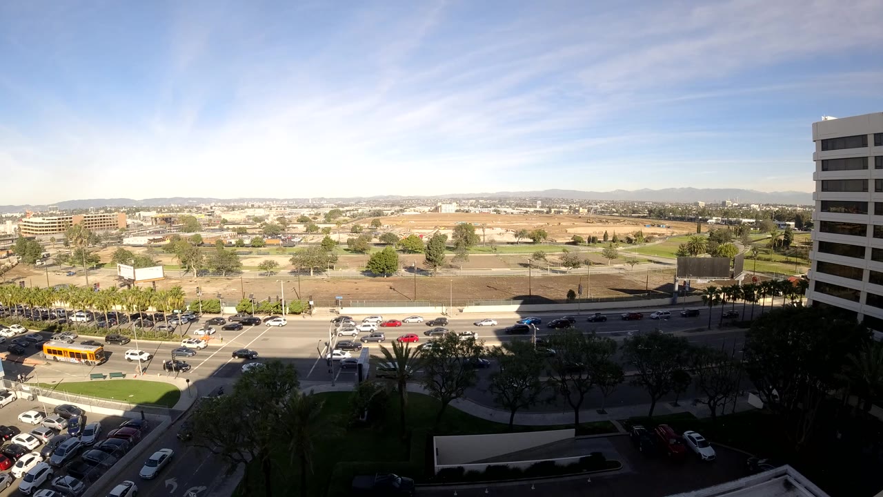 Elevated daytime view of a road, moving vehicles, lawns, trees, and distant hills.