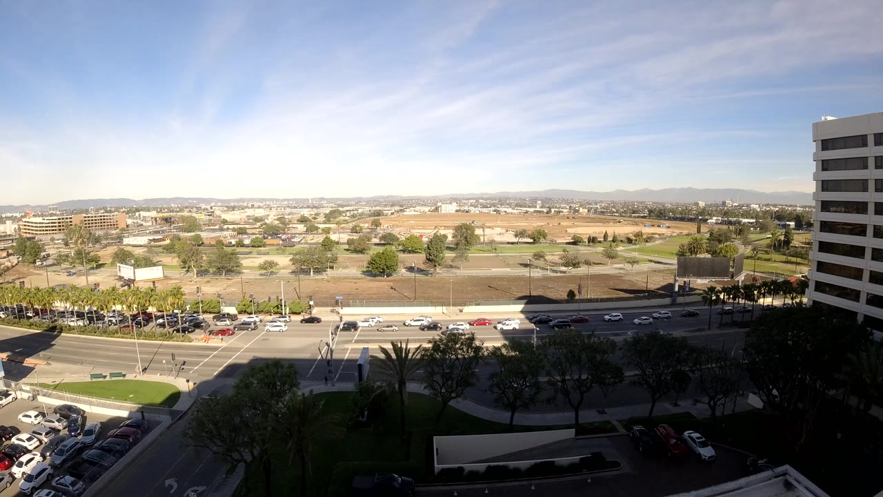 Opening frame of Los Angeles in daylight with multi-lane traffic, parking lots, trees, distant hills, and a tall office building on the right, with no visible people.