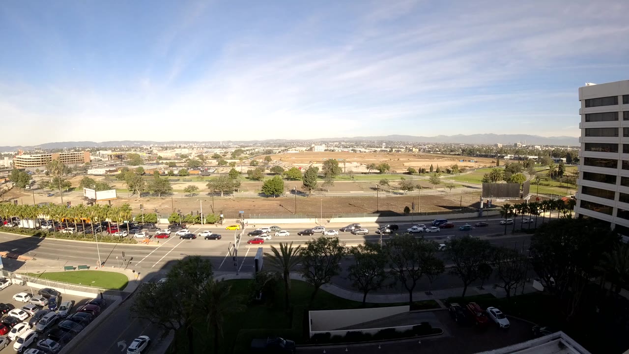 Opening frame of Los Angeles traffic seen from a high vantage point, with cars on a multi-lane road, parking areas, distant hills, and a tall white building at right with no visible people.
