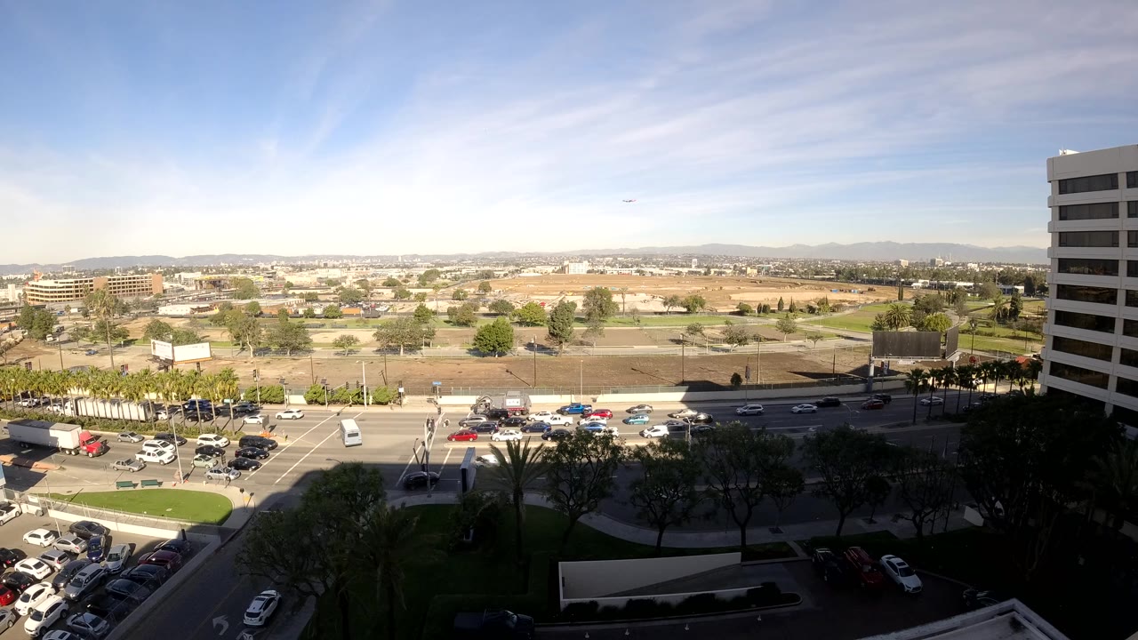 A high Los Angeles view shows cars, a white van or truck, and traffic signals near an intersection, with open lots, low-rise buildings, hills, and no visible people.