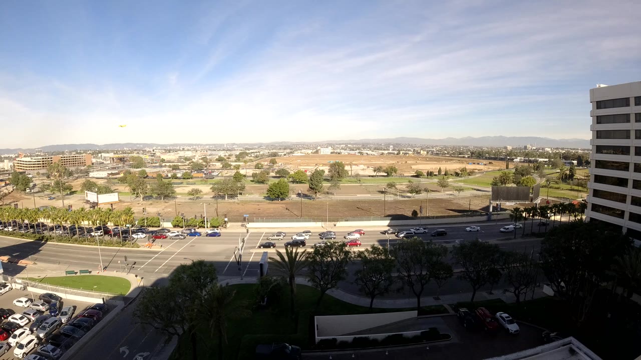 At 0:01, Los Angeles traffic moves through the intersection while a tall office building frames the right side and open land stretches toward distant hills, with no visible people.