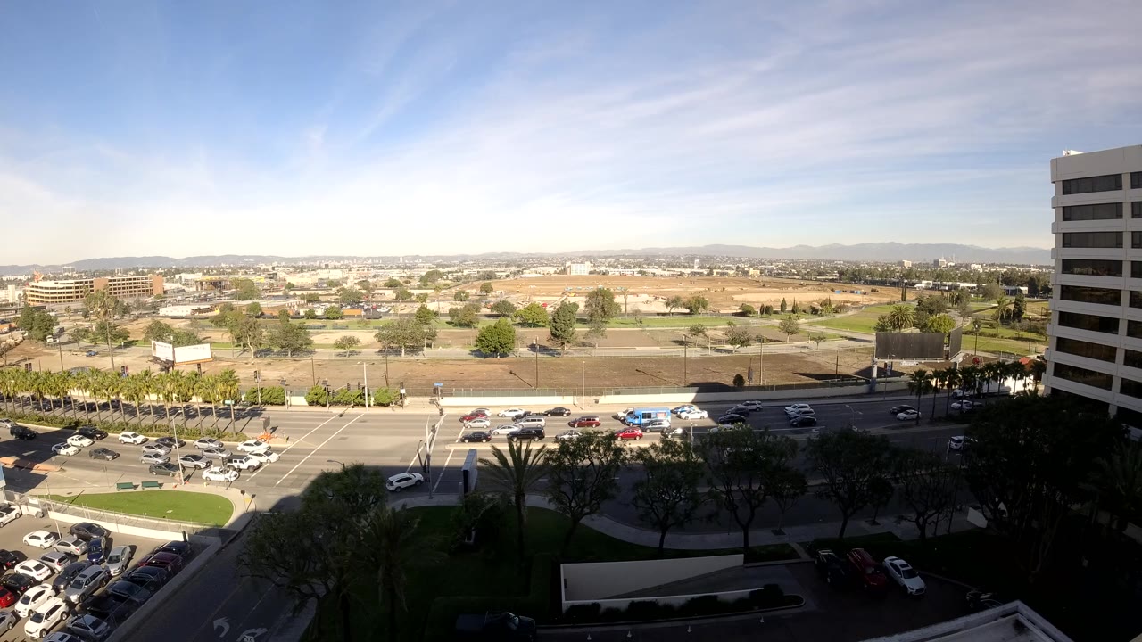 At 0:02, cars and light-colored vehicles cluster around the Los Angeles intersection, with parking lots below, low buildings beyond, and no visible people.