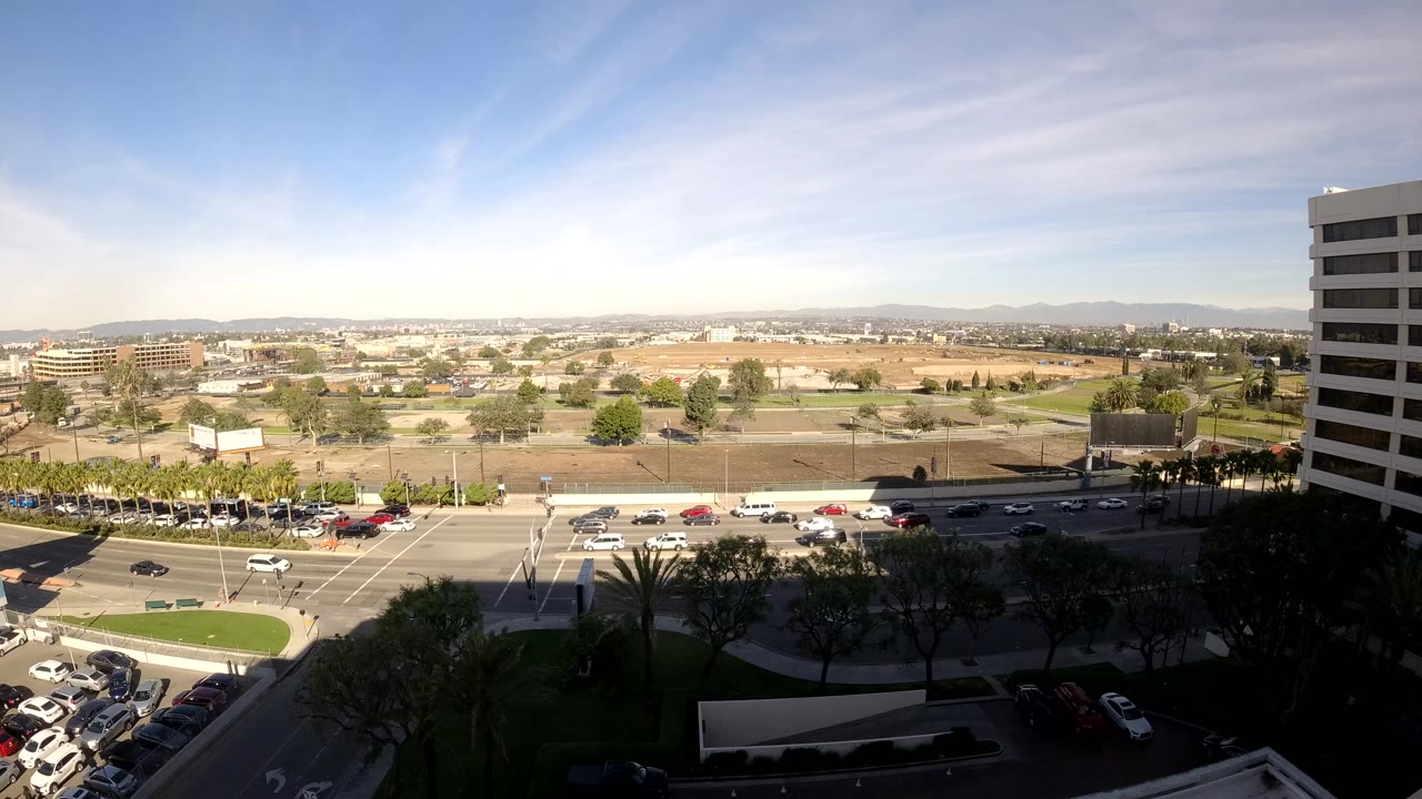 Wide daytime city view with traffic, parking lots, trees, and mountains beyond.