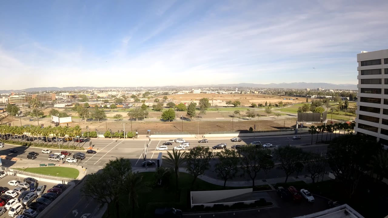 Los Angeles, California high daytime frame with a multilane intersection, several cars, a bus or shuttle visible near the road, office buildings, trees, distant mountains, and no visible people.