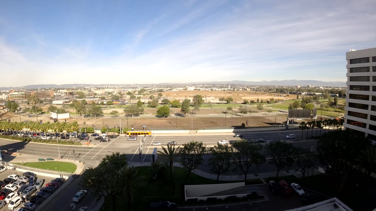 High daytime view over a Los Angeles intersection with cars on multiple roads, parking lots at lower left, a white office tower at right, distant hills, and no visible people.