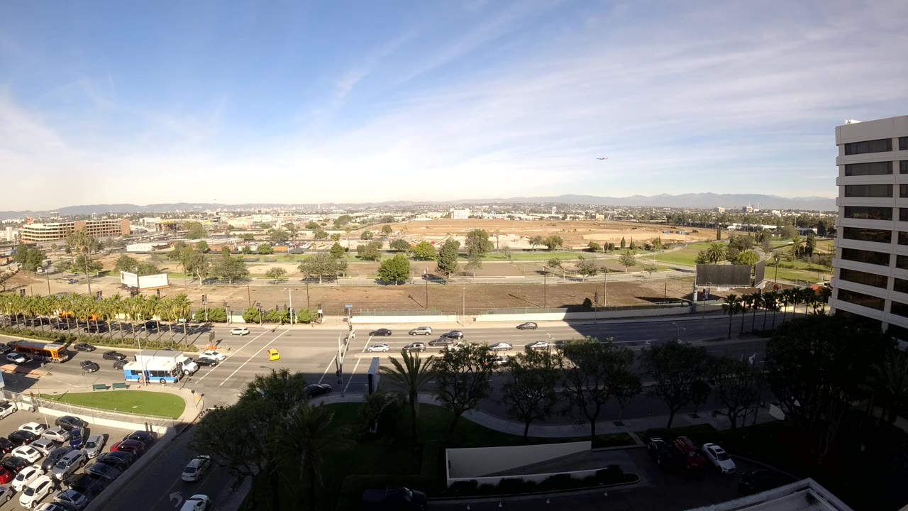 Los Angeles traffic scene from above with cars and buses near the intersection, bright road surfaces, a right-side office tower, distant low-rise development, and no visible people.