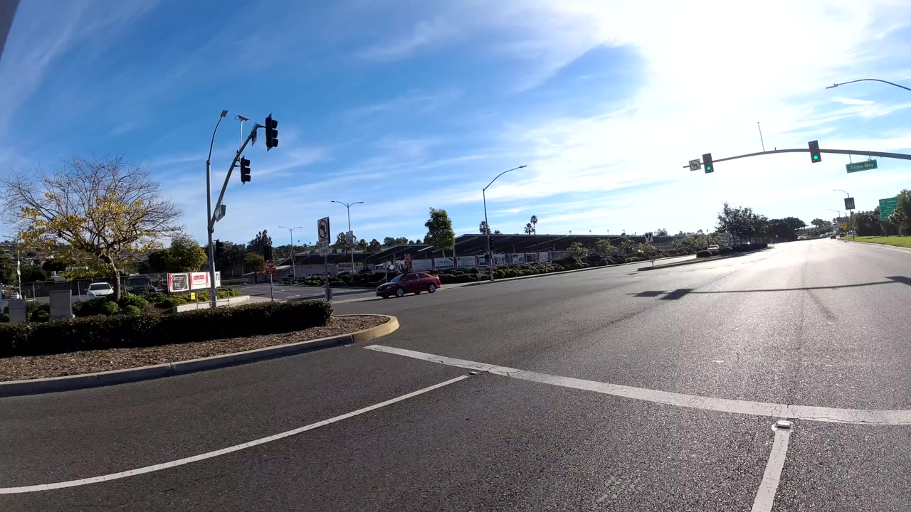 Wide vehicle-height view of an Orange, California road intersection with traffic signals, painted lane lines, roadside trees, low buildings, blue sky, sun glare, and no visible people.