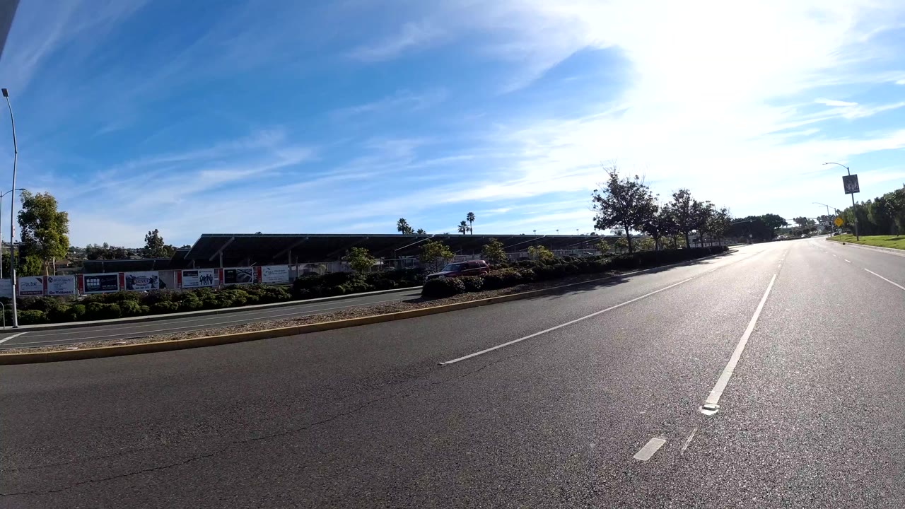 Forward road view with empty lanes, median planting, roadside buildings, trees, and bright sky.