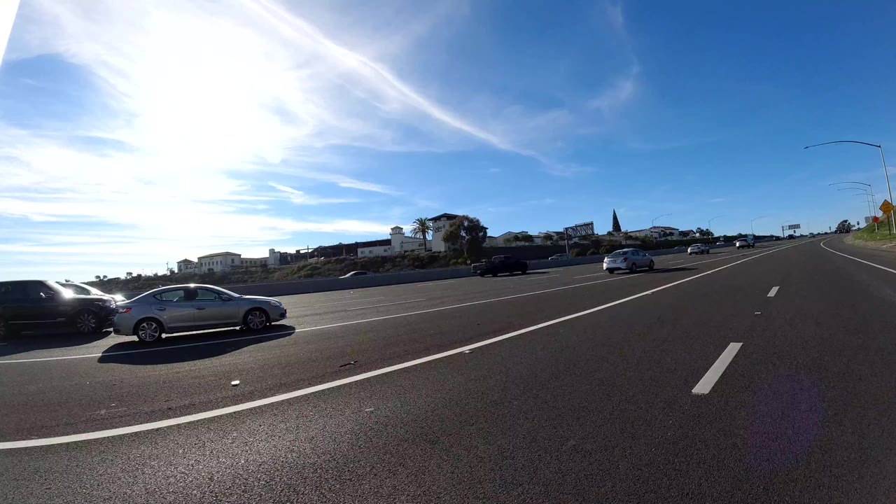 Side and forward freeway view in Orange, California with two cars near the camera, multiple lanes, low roadside buildings, bright blue sky, sun glare, and no visible people.