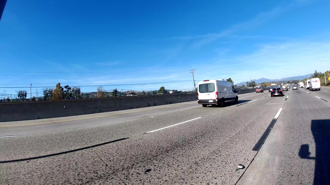 A white van and several cars travel along a multi-lane freeway in Orange, California, with concrete barriers, distant hills, and no visible people.