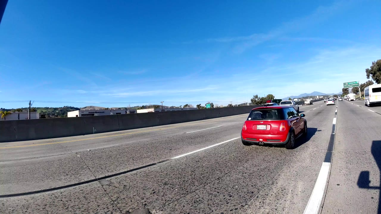 A red hatchback passes near the camera on the freeway in Orange, California, with other cars, roadside signs, distant hills, and no visible people.