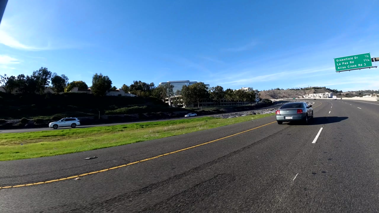 Open Orange, California freeway lane with a green overhead sign, palm tree, dark roadside trees, cars in the distance, strong sun, and no visible people.