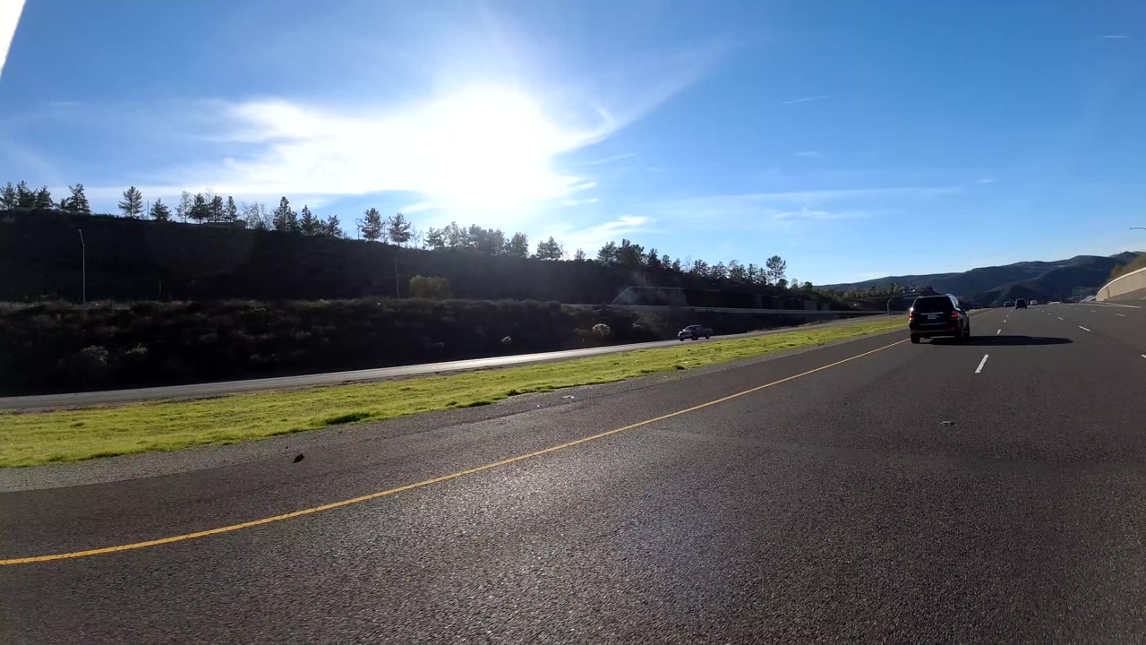 Cars on a sunny freeway with green shoulder and hillside in the distance