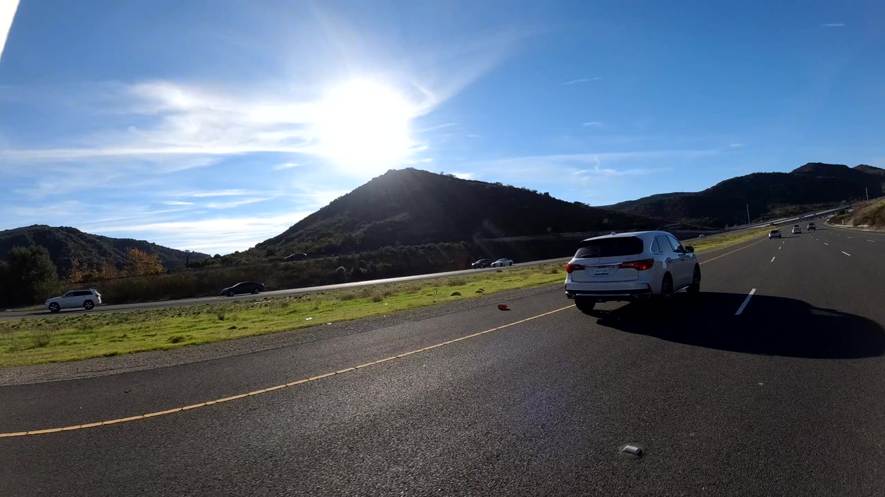 A white SUV passes on the right side of a freeway in Orange, California, with a sunlit hill, green roadside, blue sky, and no visible people.