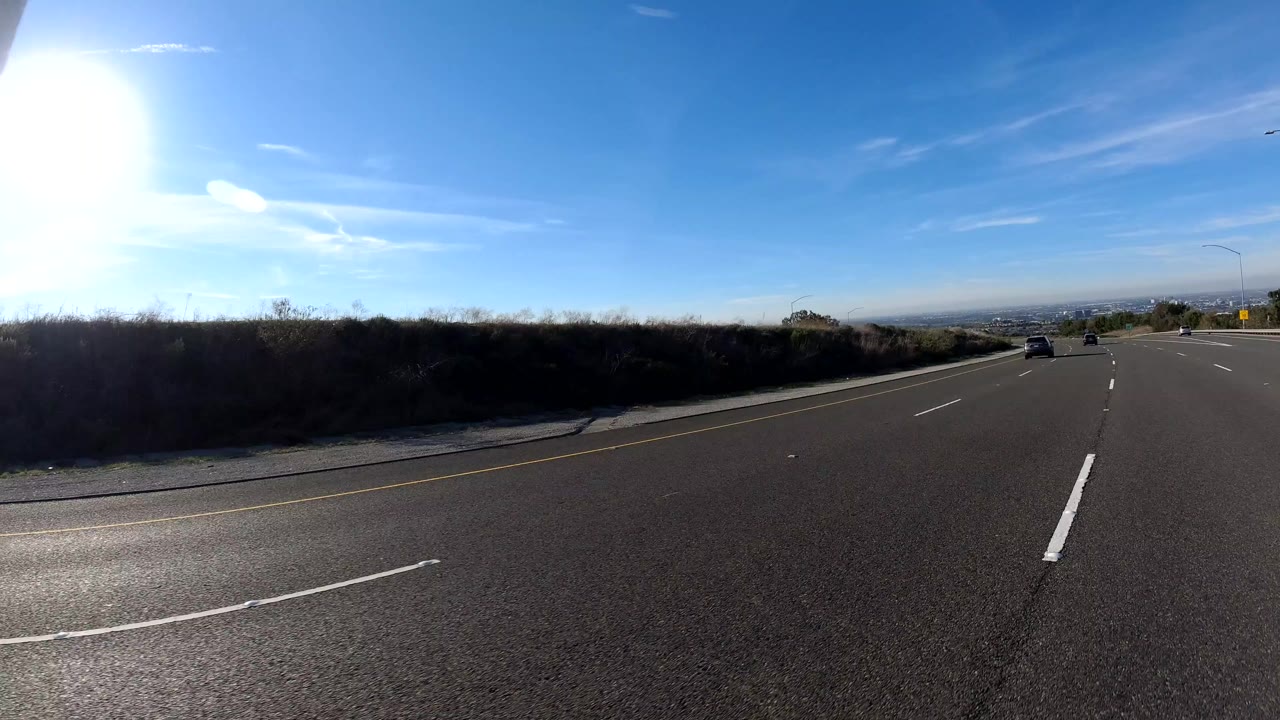 Orange, California freeway lanes run past dark roadside brush with a bright sun flare on the left, a clear blue sky, and no visible people.