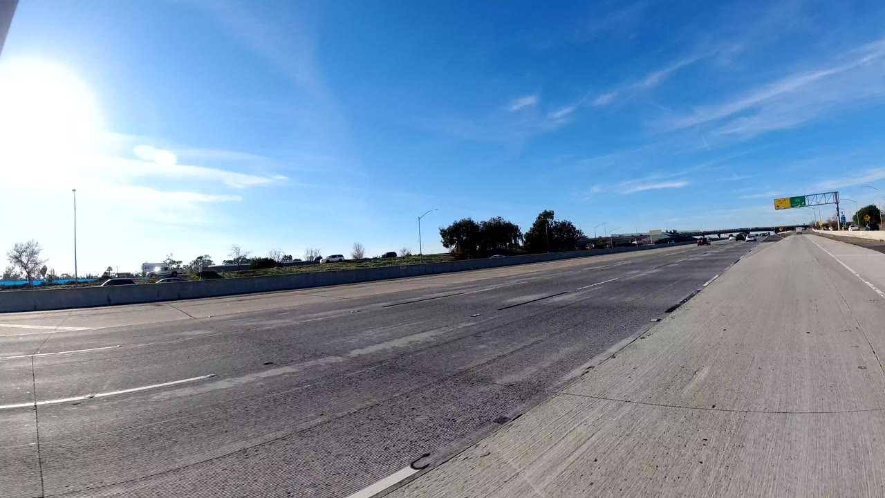 Wide road-level view of freeway lanes in Orange, California, with bright sun at the left edge, blue sky, distant cars, roadside trees, and no visible people.