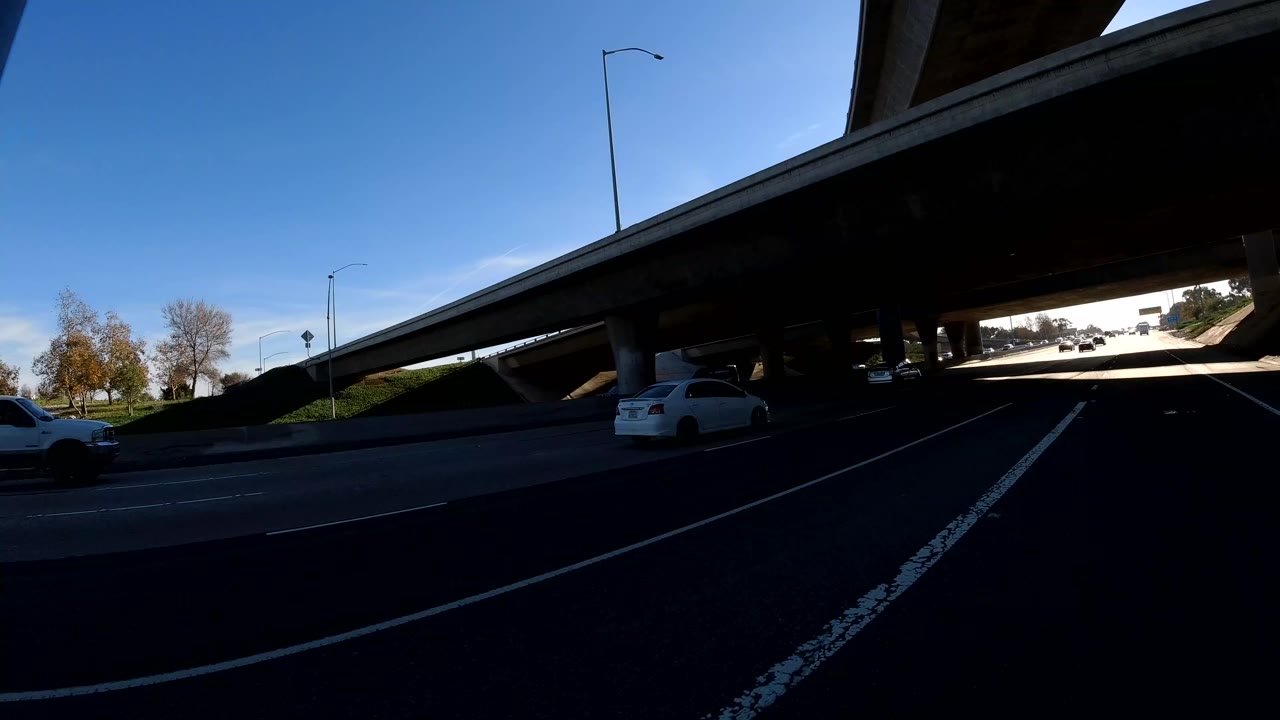 Dark underpass view in Orange, California, showing roadway lanes, cars ahead and to the side, overpass columns, ramp structure, and no visible people.