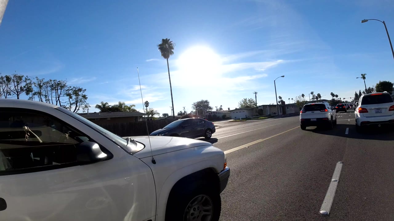 Orange, California roadway scene with a white vehicle close at left, cars ahead in traffic lanes, palm trees, streetlights, bright sun glare, and no visible people.