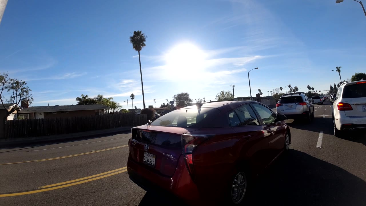Orange, California roadway frame with a red car close in the right foreground, several cars ahead, palm trees, streetlights, blue sky, and no visible people.