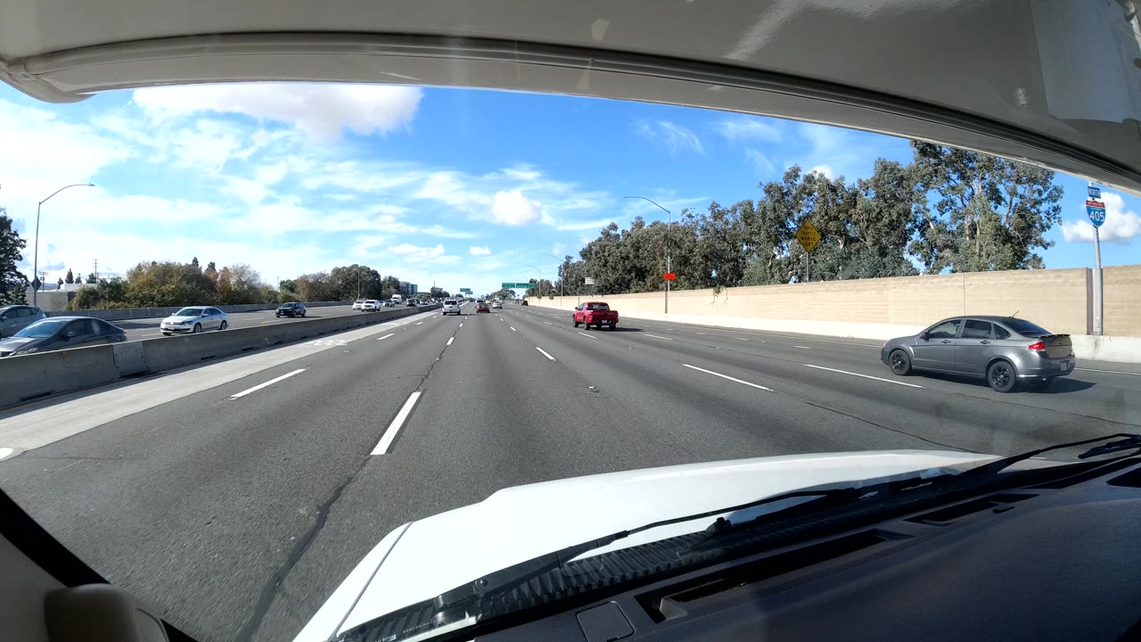 Opening California coast freeway view from behind a windshield, with the vehicle hood in front, traffic spread across several lanes, a wall and trees on the right, and no visible people.