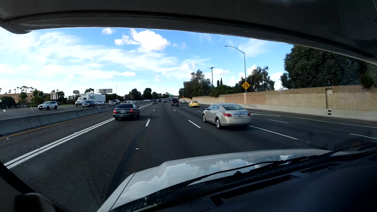 The dashboard-like view shows cars ahead and to the right on a California coast freeway, with a tan roadside wall, trees, open blue sky, and no visible people.