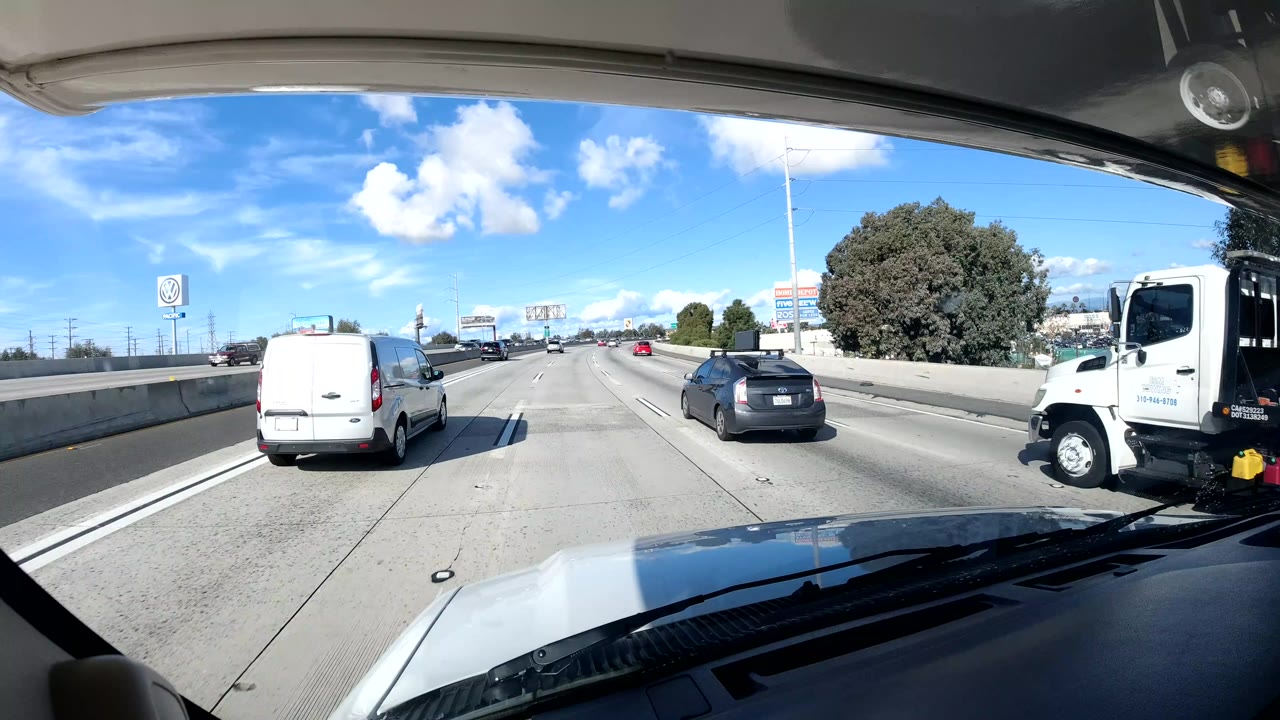 Dashboard view on a California coast freeway with a white van, nearby cars, concrete lanes, roadside trees, bright sky, and no visible people.